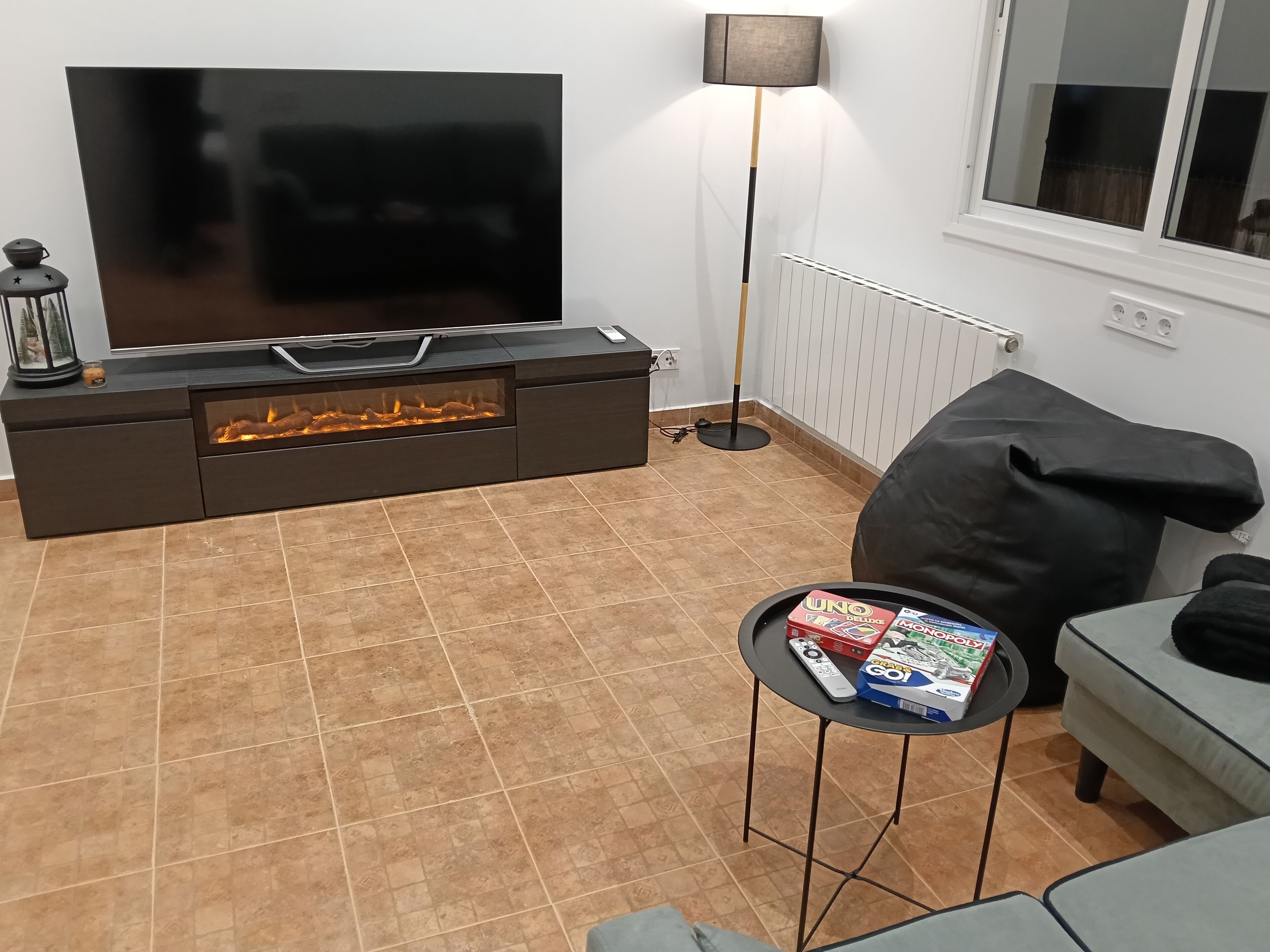 A living room with a flat-screen TV, a fireplace, a standing lamp, a sofa, and a small table with board games.