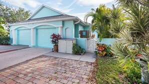 A light blue house with two garage doors, a brick driveway, and a small garden with various plants. - Coastal Villa, Walk to the Beach (Cape Canaveral)