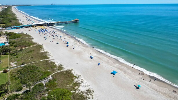 An aerial view of a beach with a pier extending into the ocean, numerous umbrellas, and a clear blue sky. - Coastal Villa, Walk to the Beach (Cape Canaveral)