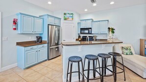A modern kitchen with blue cabinets, a stainless steel refrigerator, a microwave, and a coffee maker. There are two stools at the island and a white door in the background. - Coastal Villa, Walk to the Beach (Cape Canaveral)