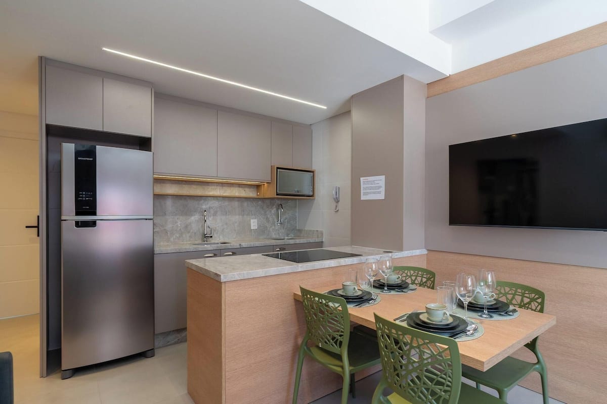 A modern kitchen with a built-in fridge, microwave, and sink. There is a dining area with a table set for two, green chairs, and a flat-screen TV mounted on the wall.