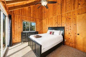 A wooden-paneled bedroom with a bed, a nightstand, a window, and a ceiling fan. - The Hidden Pines Mountain Home by Navigate Stays (Running Springs)