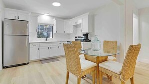 A modern kitchen with white cabinets, a stainless steel refrigerator, and a glass dining table with wicker chairs. - Cottage on California (Stuart)