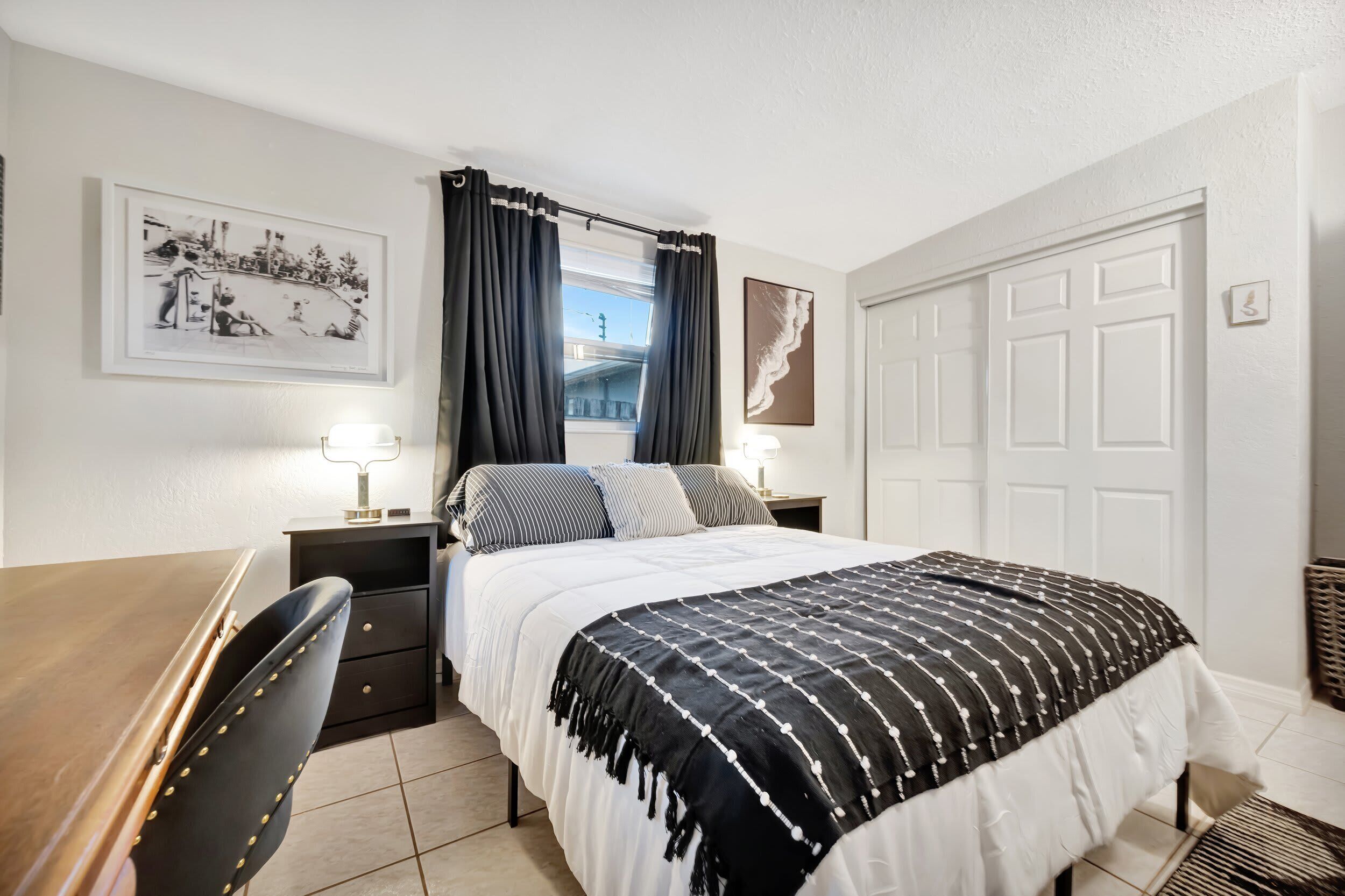 A bedroom with a bed, a desk, a chair, a nightstand, and a window with curtains.