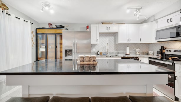 Een moderne keuken met witte kastjes, een groot kookeiland, roestvrijstalen apparatuur en een backsplash met een visgraatmotief.