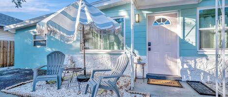 A house with a number 5025, a porch with two chairs, a striped umbrella, and a welcome mat.
