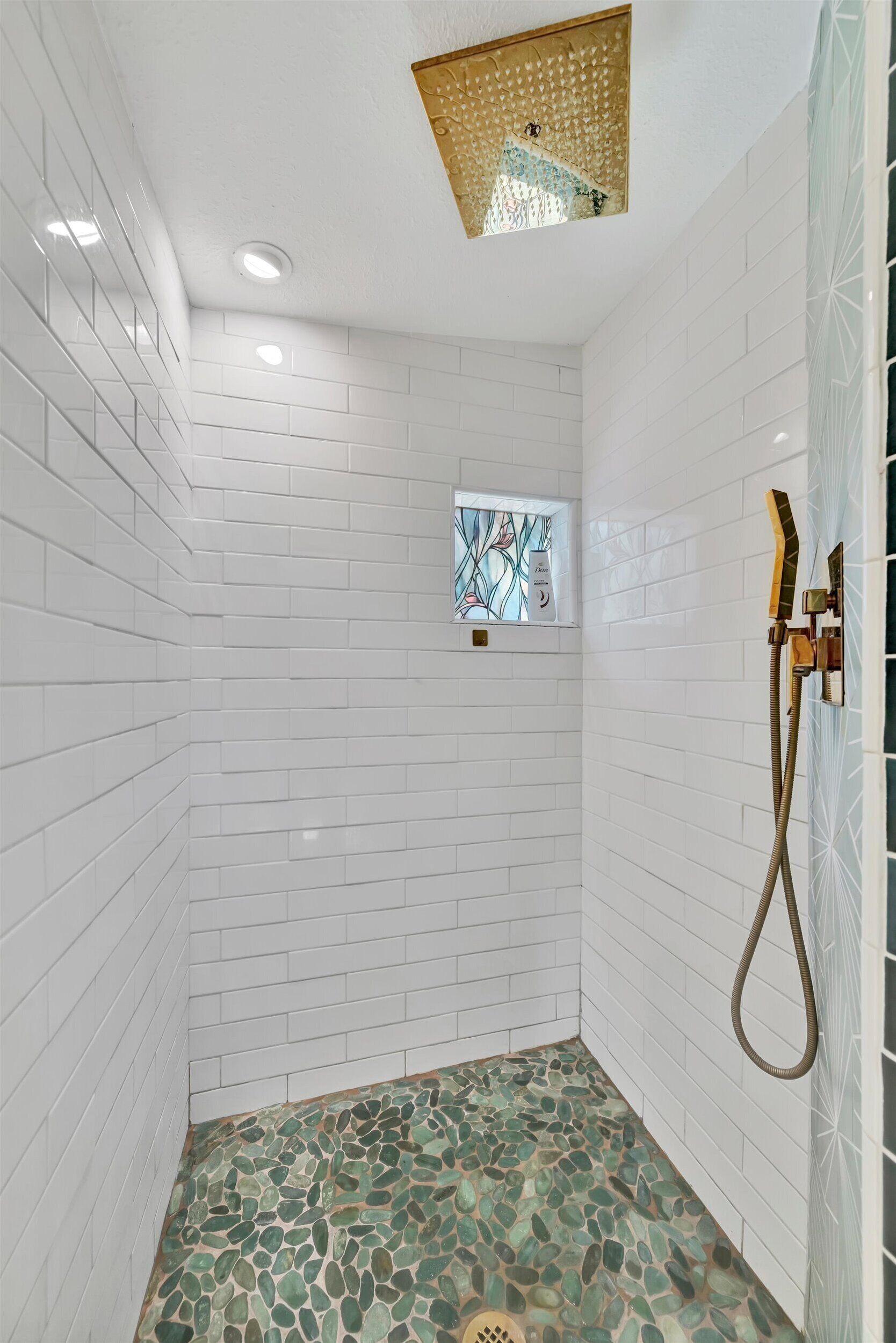 A shower with white tiled walls, a mosaic floor, and a small window with stained glass.
