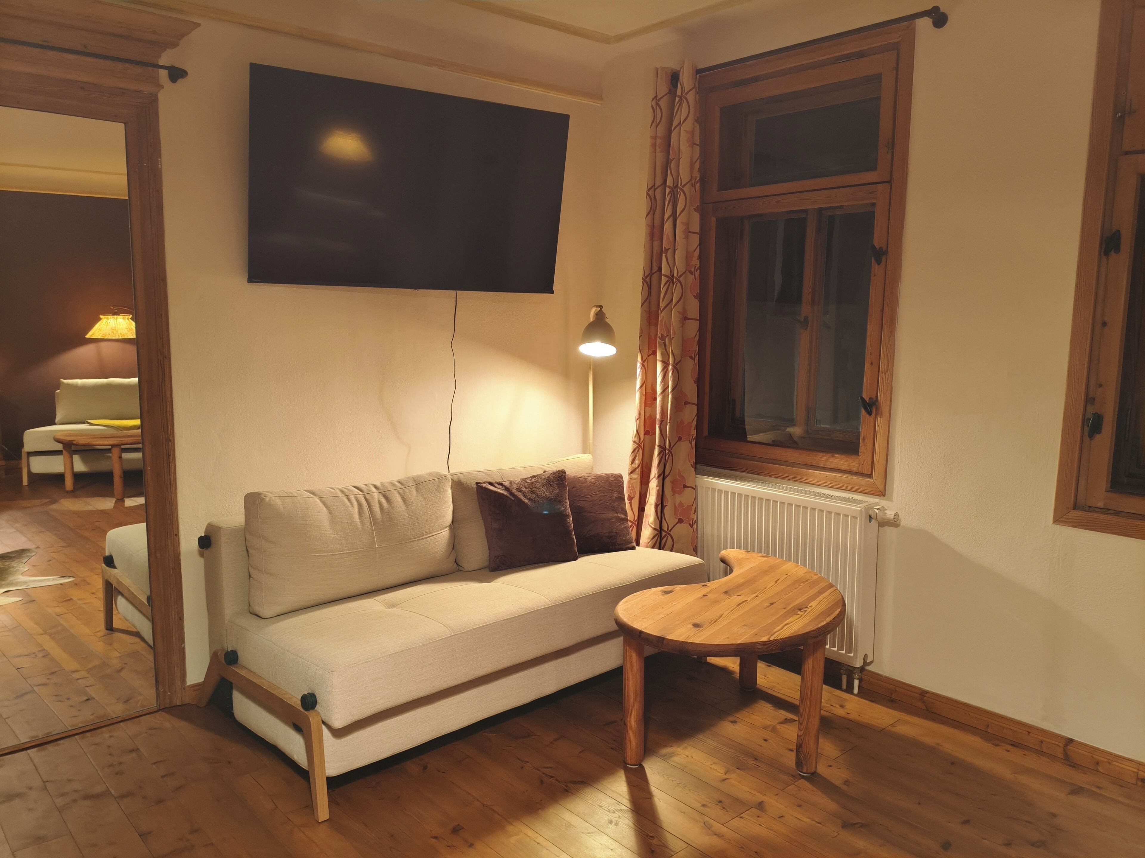 A living room with a sofa, a round wooden table, a TV mounted on the wall, and a window with curtains.