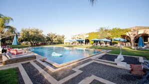 Una piscina con sillas de descanso y sombrillas, rodeada de un jardín ajardinado y un edificio al fondo.