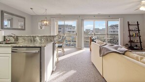 Ett modernt kök med diskmaskin, en matplats med stolar, en soffa och utsikt över havet genom stora skjutdörrar.