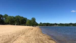 A sandy beach with a clear blue sky, trees, and a calm lake. - Lakeside Hideaway (Lake Ronkonkoma)