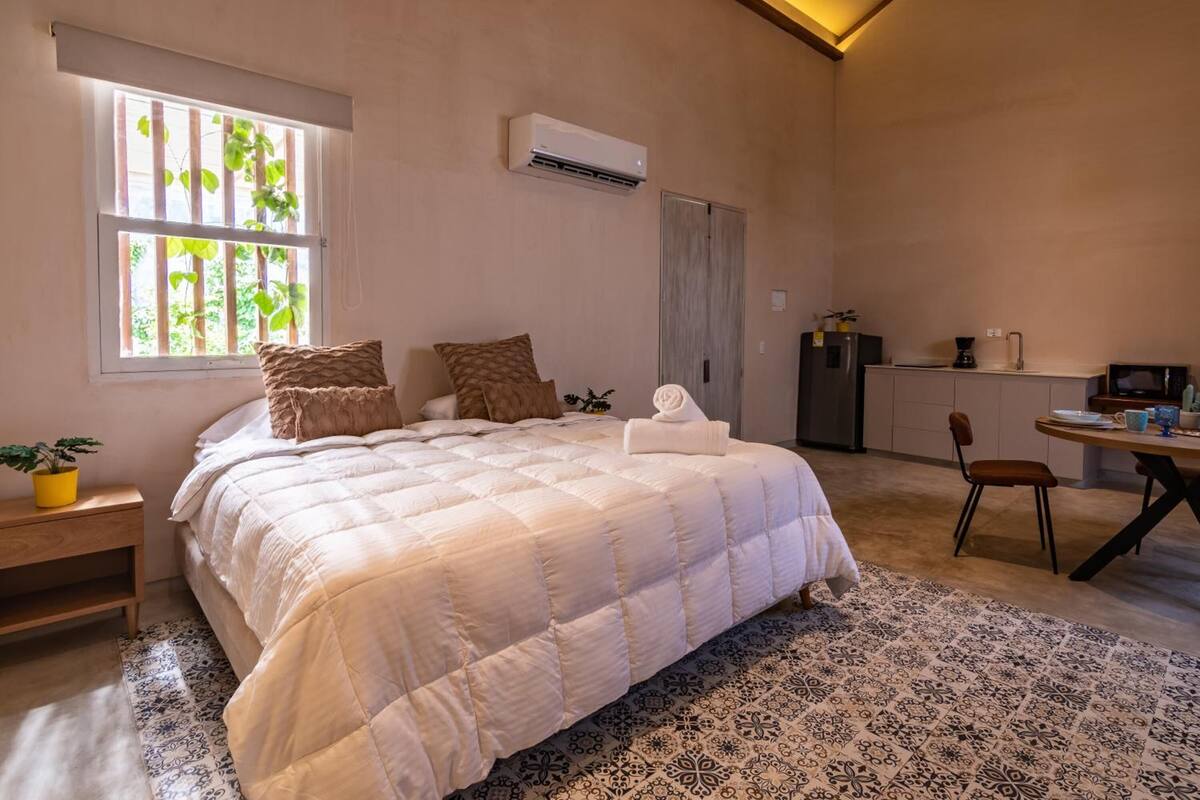 Un dormitorio con cama, almohadas, una mesita de noche con planta, ventana con persianas, equipo de aire acondicionado, kitchenette con microondas y una silla.