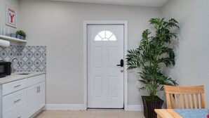 Eine kompakte Küche mit weißen Schränken, einer Mikrowelle und einer gemusterten Spritzschutzwand. Eine weiße Tür mit rundem Fenster ist zu sehen, und rechts daneben steht ein Blumentopf.