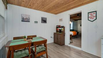 A cozy room with a wooden ceiling, a dining table with chairs, a small cabinet, and a bed in the background.