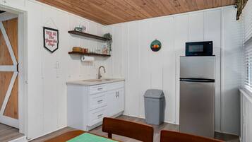 A compact kitchen with white cabinetry, a stainless steel refrigerator, a microwave, and a trash bin. There is a wooden door on the left and a small shelf above the sink.
