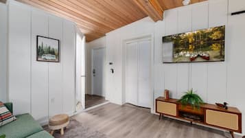 A spacious living room with a wooden ceiling, a green sofa, a small rug, a wooden console table with a plant, and a painting of boats on a lake.