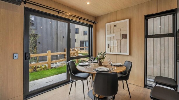 A modern dining area with a round table, black chairs, and a view of a wooden deck and building outside. - Remarkable Escape | Modern Central Stay Near Airport (Queenstown)