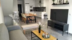 Een moderne woonkamer met een bank, een salontafel en een televisie. Er is een eethoek met een houten tafel en stoelen.