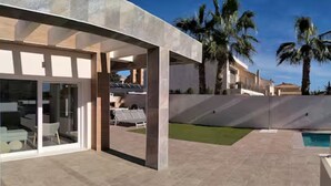 Een modern huis met een terras, een zwembad en palmbomen.