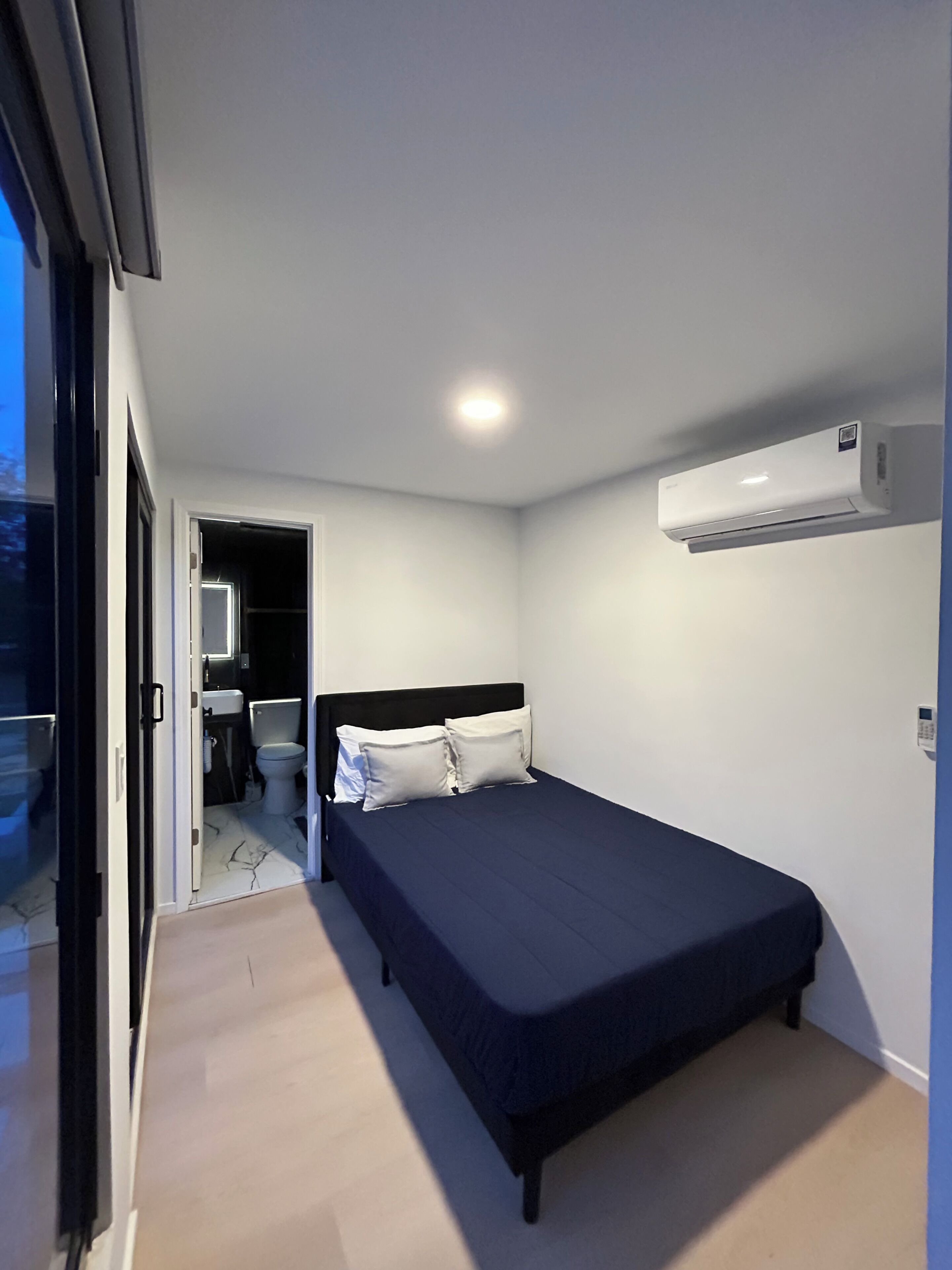 A bedroom with a bed, a wall-mounted air conditioner, and a bathroom visible through an open door.