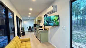 Ein modernes Wohnzimmer mit einem gelben Sofa, einem Kuhfellteppich, einer Küche mit Fernseher und einem großen Fenster mit Blick auf einen Wald.