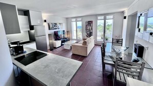 Et moderne køkken med en central ø, et spiseområde med et glasbord og en stue med sofa og fjernsyn.
