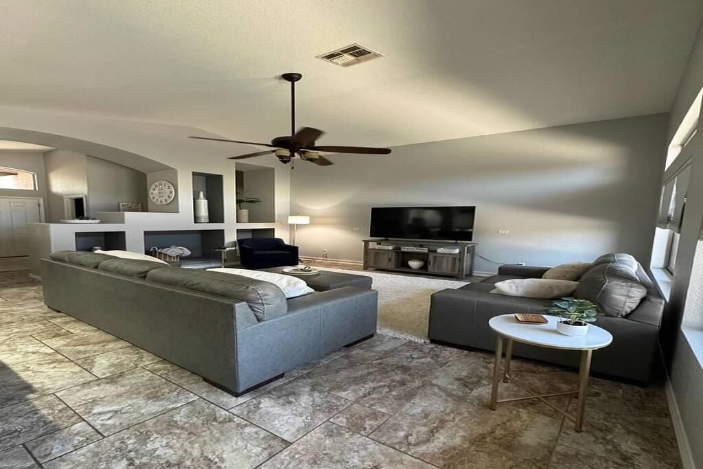 A spacious living room with a large grey sectional sofa, a smaller grey sofa, and a round coffee table. There is a flat-screen TV mounted on the wall, a ceiling fan, and a small side table with a plant.