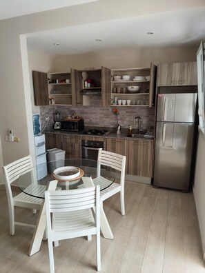 Eine moderne Küche mit Holzschränken, einem Edelstahlkühlschrank, einem Glastisch mit weißen Stühlen und einem Wasserspender.