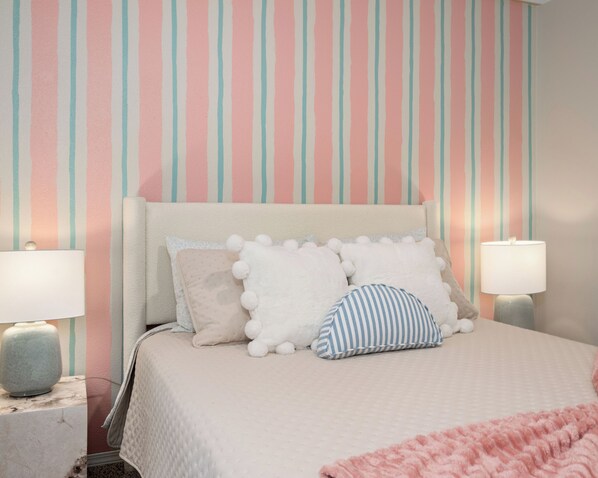 A neatly made bed with a pink blanket and three pillows, a bedside lamp, and a striped wall in pastel colors.