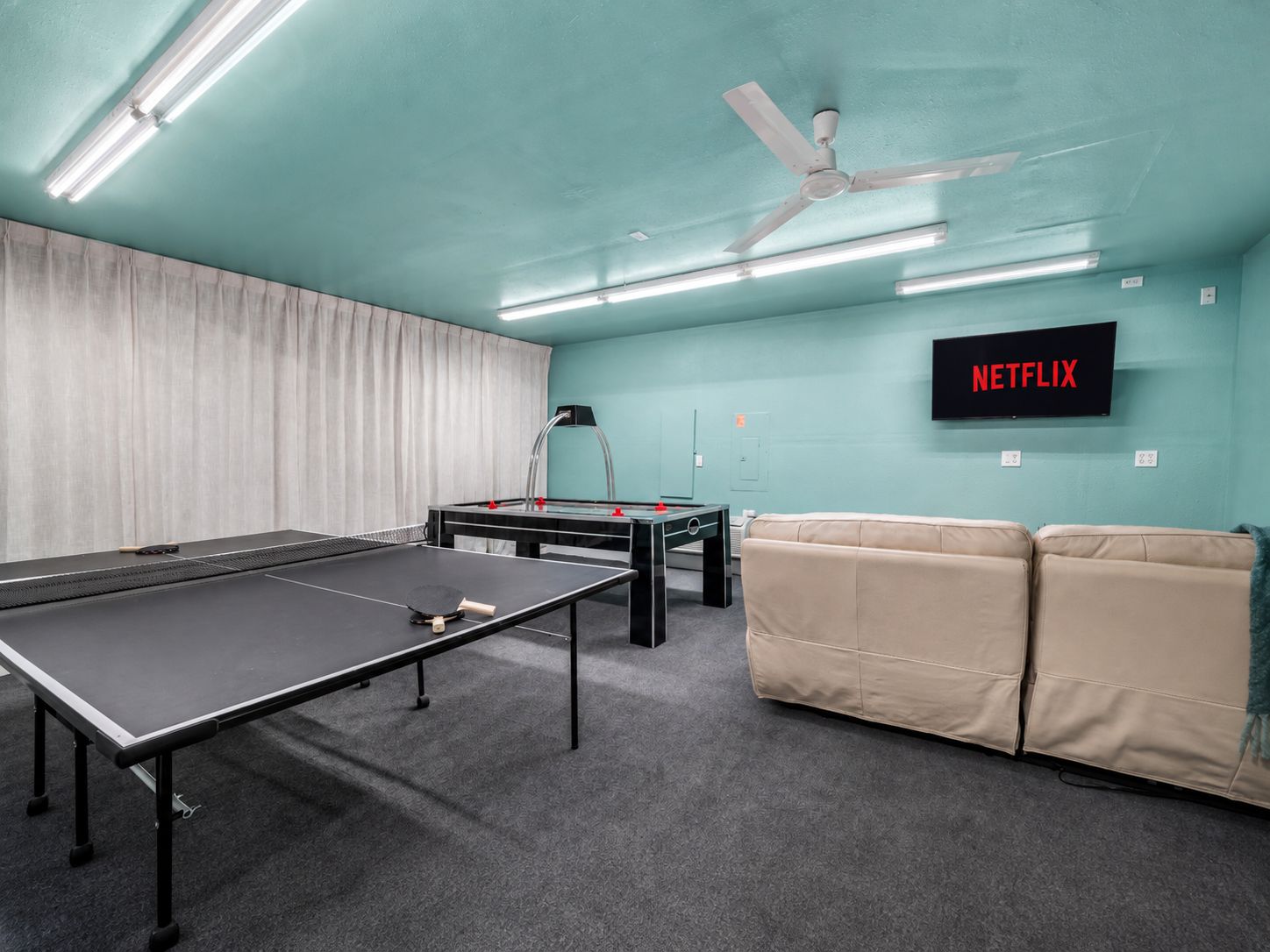 A room with a ping pong table, a pool table, a ceiling fan, and a wall-mounted television labeled 'NETFLIX'.