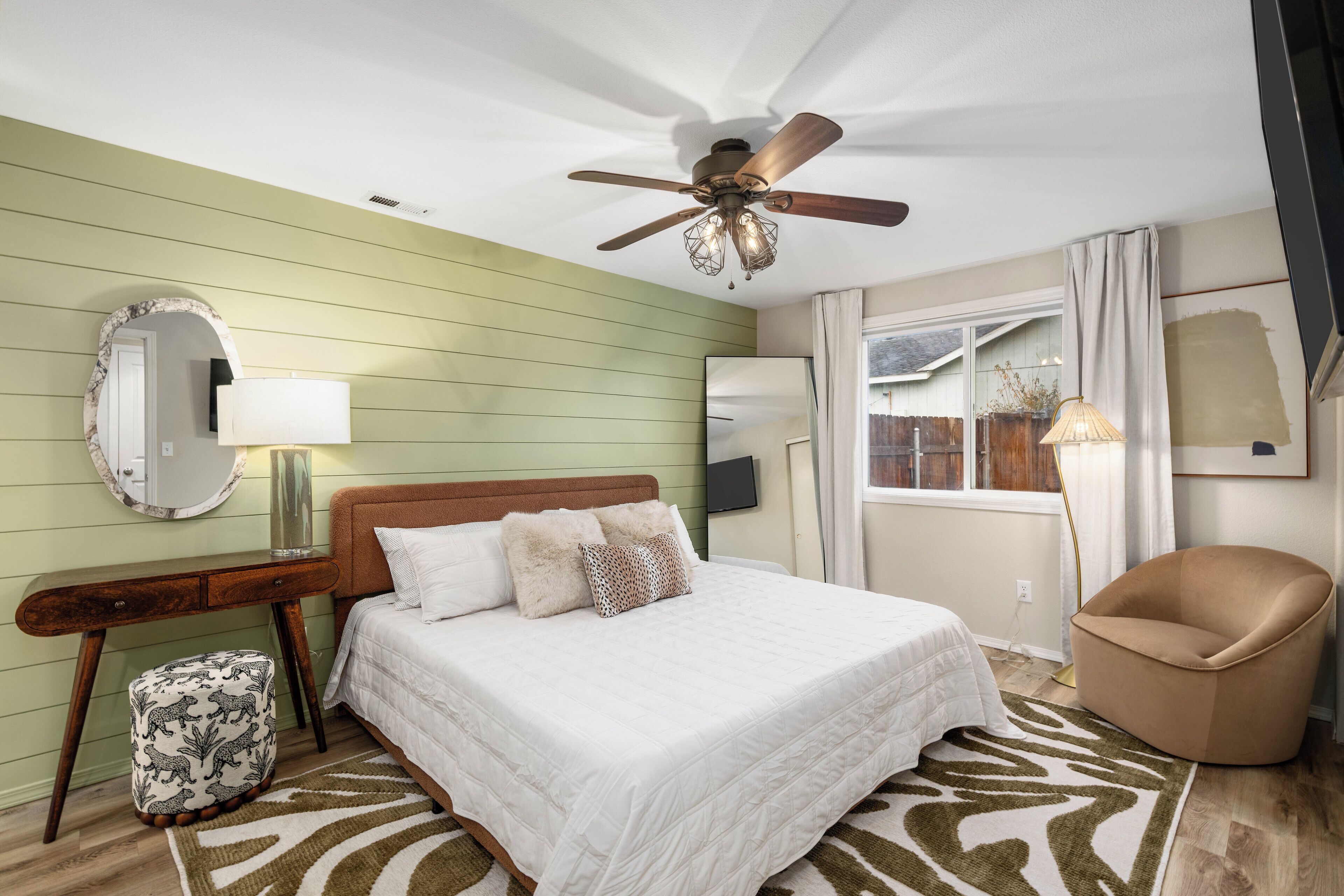 Une chambre à coucher comprenant un lit, un ventilateur de plafond, une table de chevet, une chaise, une fenêtre et un miroir.