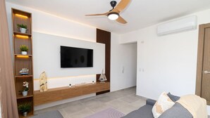 Ein modernes Wohnzimmer mit einem flachbildfernseher an der Wand, einem Holzkonsolentisch mit Dekoration, einem grauen Sofa und einem Deckenventilator.