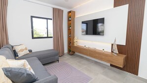 Ein modernes Wohnzimmer mit grauem Sofa, einem Flachbildfernseher, einem Holztischchen und Blick durch ein Fenster.