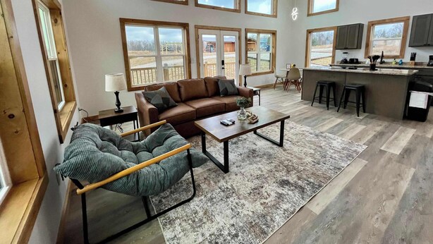 Ein geräumiges Wohnzimmer mit einer braunen Ledersofa, einem grünen Sessel und einem Couchtisch. Mehrere Fenster sorgen für natürliches Licht und bieten einen Ausblick ins Freie.