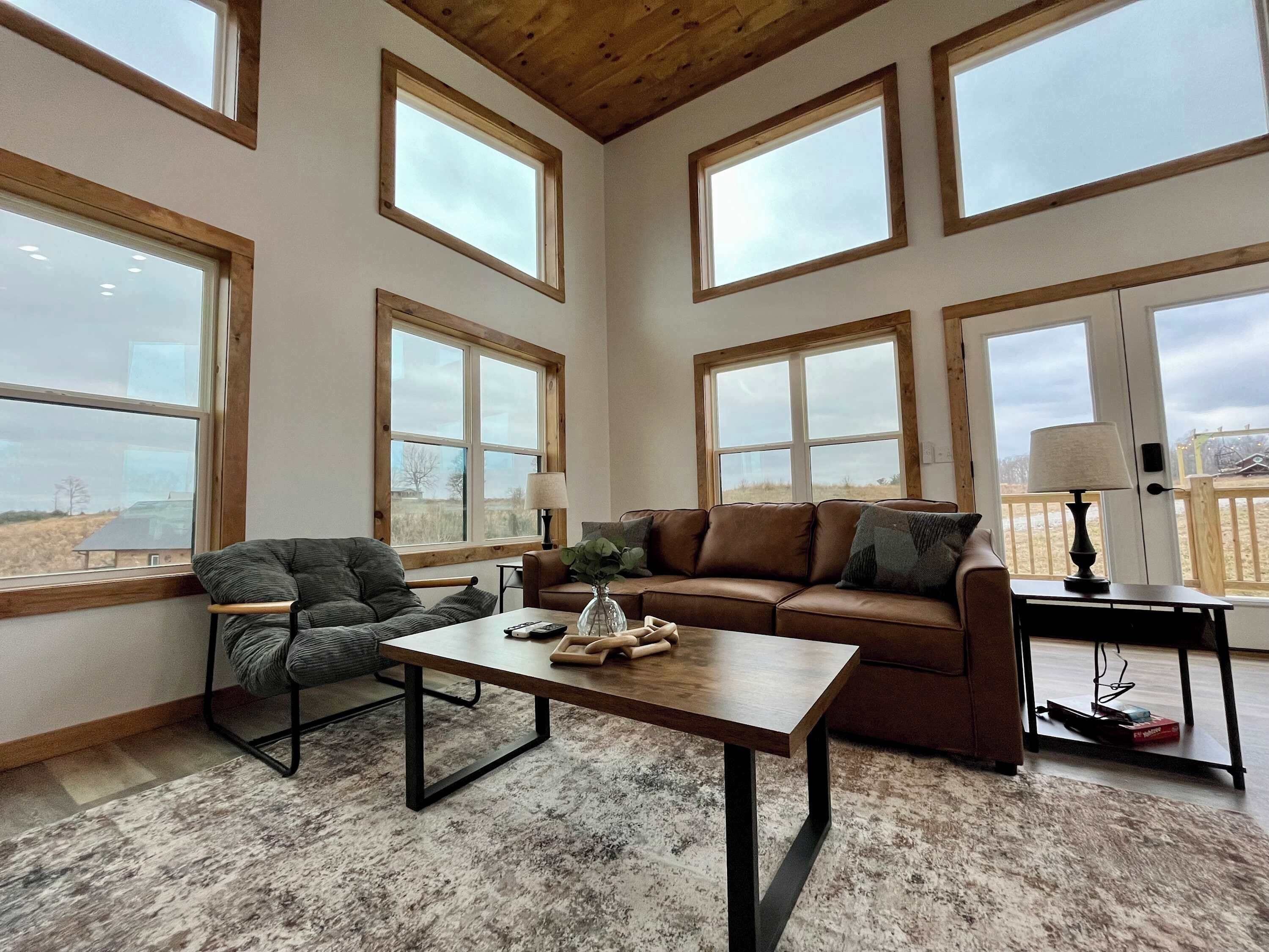 A spacious living room with large windows, a brown leather sofa, a wooden coffee table, and a chair.