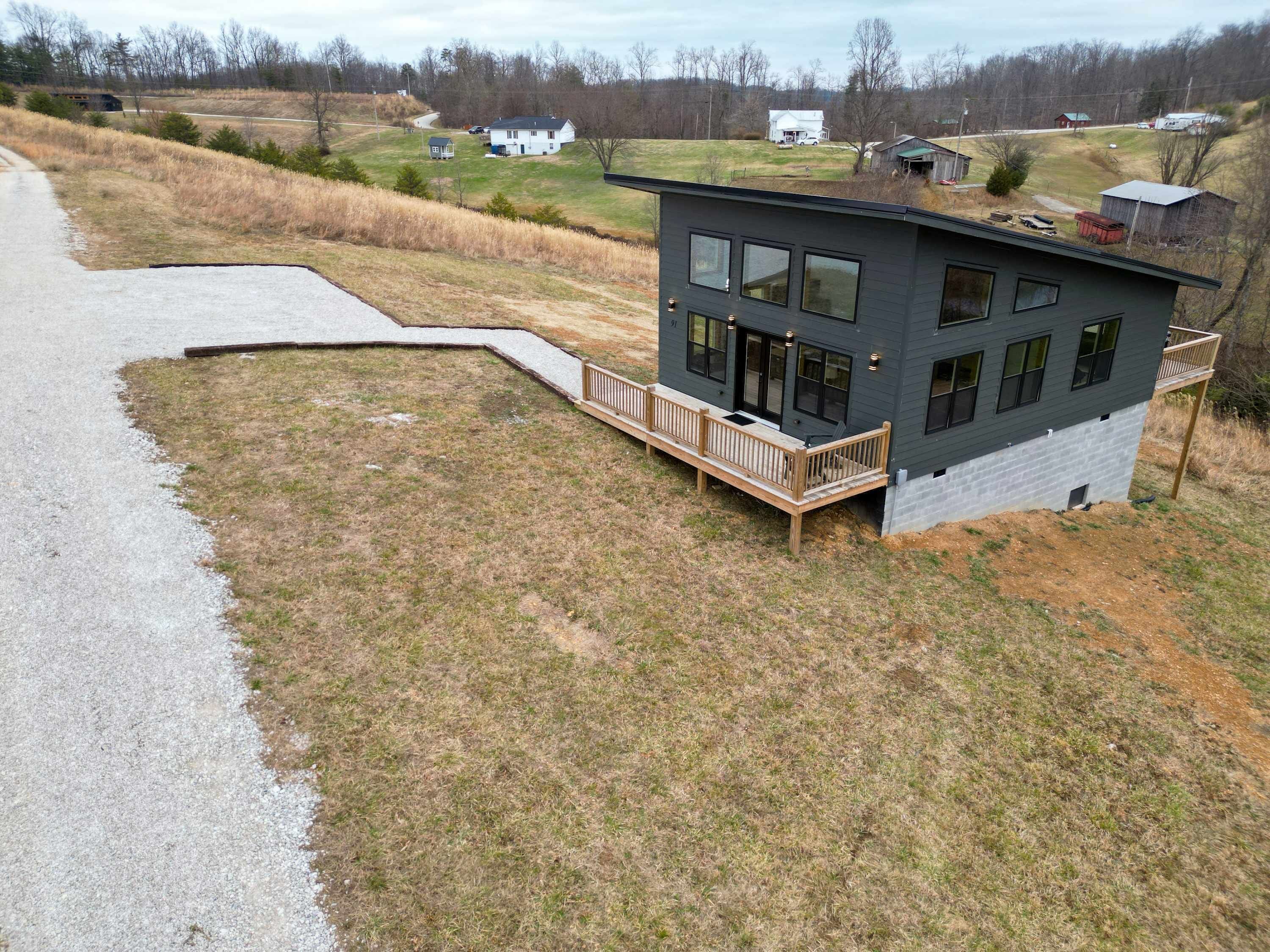 A modern two-story house with a deck, surrounded by a grassy field and a paved driveway.