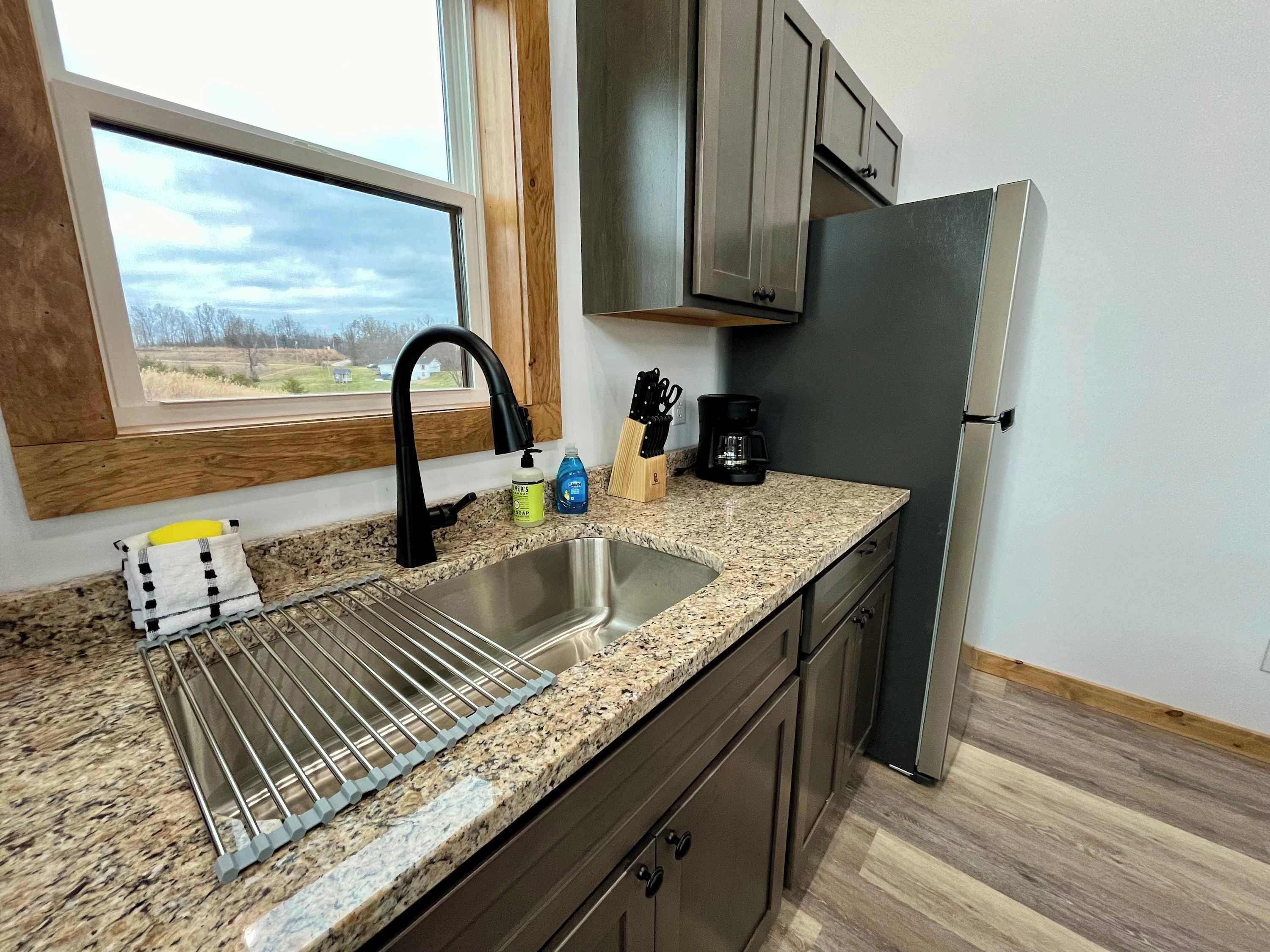 A kitchen with granite countertops, a stainless steel sink, a window with a view, and a refrigerator.