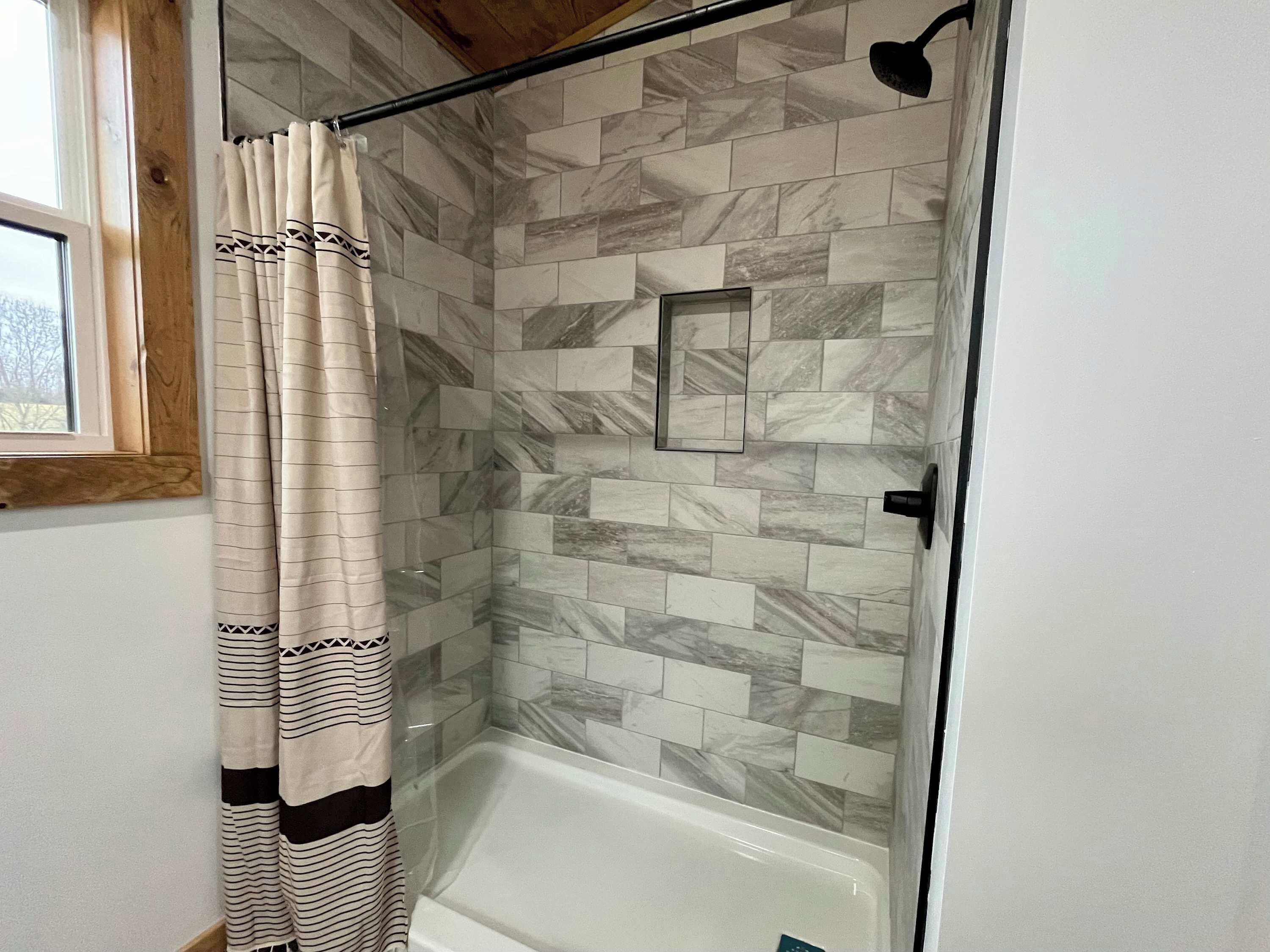 A shower with a glass door, a tiled wall, and a window with a wooden frame.