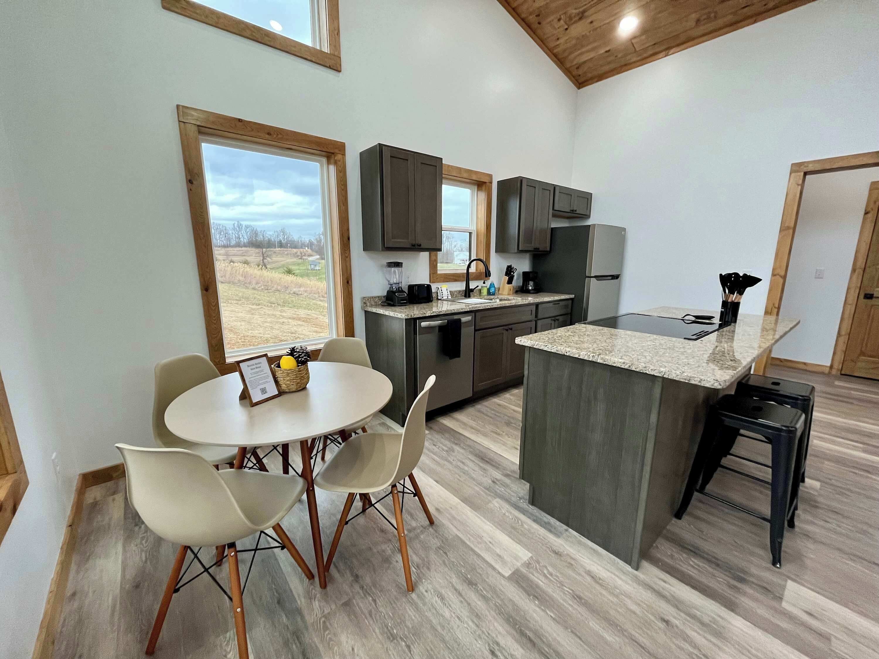 Een moderne keuken met een centraal eiland, twee eetkamerstoelen, een tafel en uitzicht op de omgeving door grote ramen.
