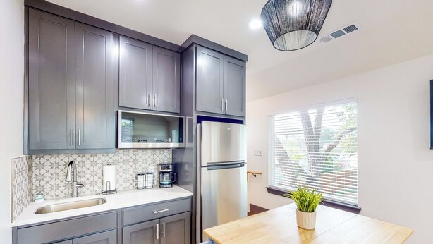 A modern kitchen with grey cabinets, a stainless steel refrigerator, a microwave, and a wooden dining table with a potted plant.