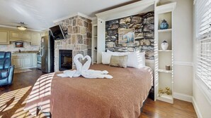 A bedroom with a stone fireplace, a bed with a brown bedspread, a TV mounted on the wall, and a kitchen in the background. - Pool + Riverfront! Couples Retreat in Smoky Mtns (Townsend)