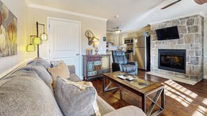 A living room with a stone fireplace, a ceiling fan, and a kitchen in the background. - Pool + Riverfront! Couples Retreat in Smoky Mtns (Townsend)