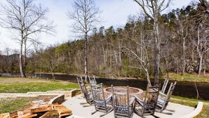 A circular outdoor seating area with metal chairs and a central fire pit, surrounded by a concrete border, set against a backdrop of trees and a body of water. - Pool + Riverfront! Couples Retreat in Smoky Mtns (Townsend)