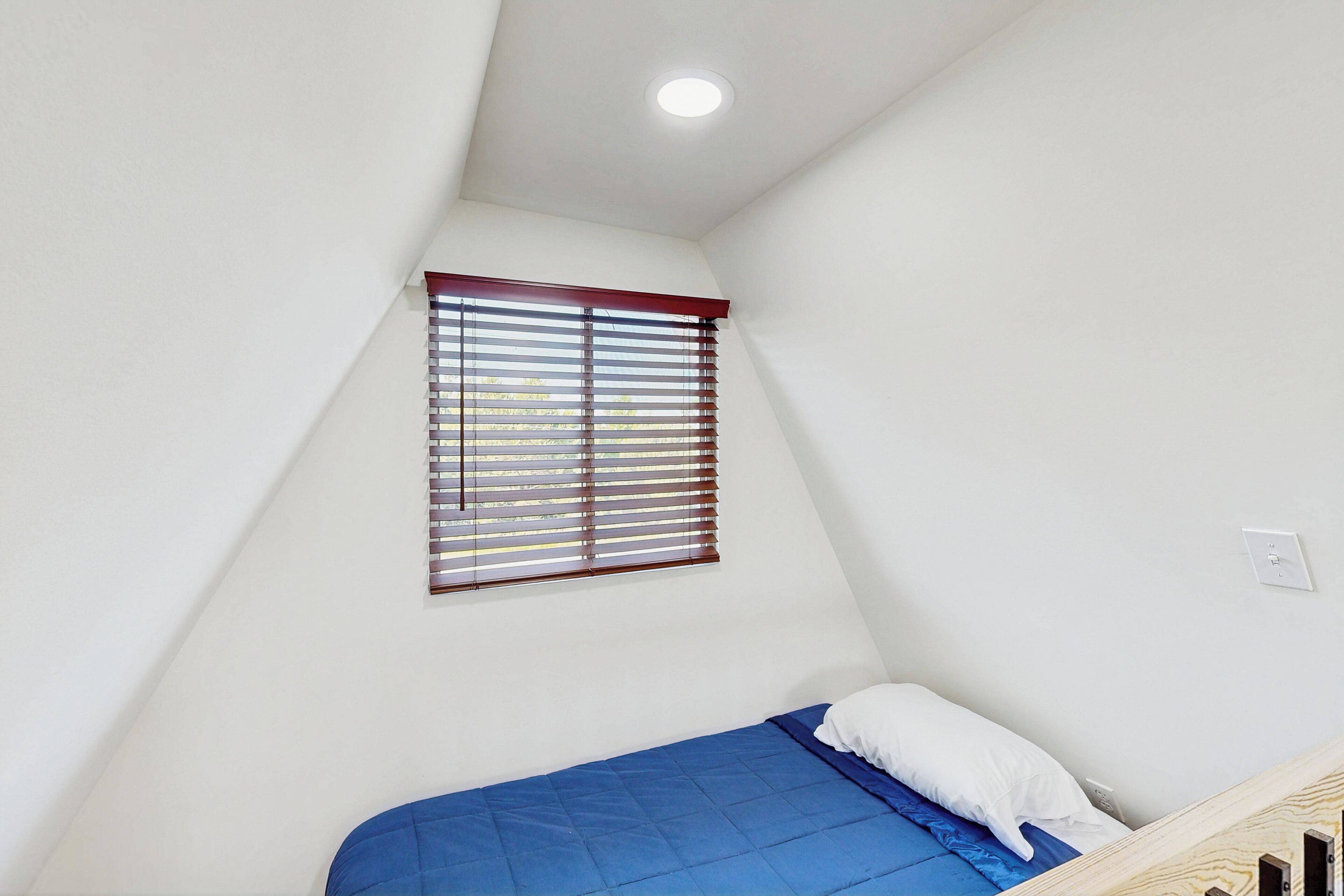 Ein kleines Zimmer mit einem Einzelbett, schrägem Dach, einem Fenster mit Jalousien und einer weißen Wand.
