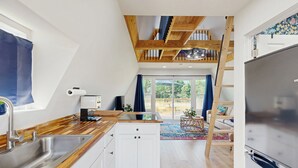 Una cocina moderna con encimeras de madera, un lavamanos de acero inoxidable y un horno empotrado. Se ve una escalera de madera que sube a un nivel superior, y un amplio ventanal con cortinas.