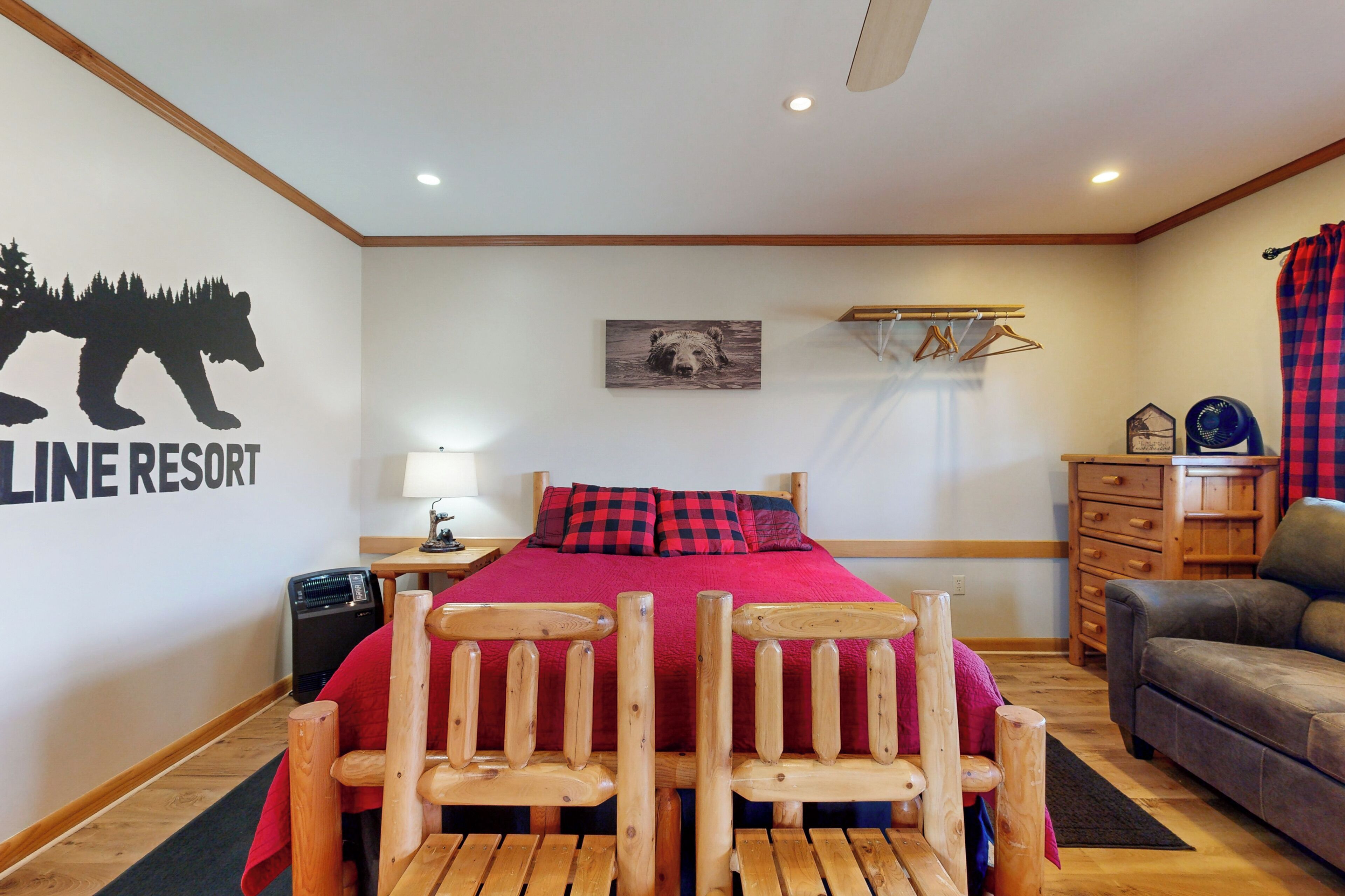 A bedroom with a bed, wooden chairs, a dresser, a lamp, and a wall with a bear silhouette and text.