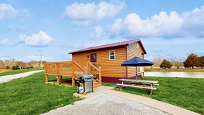 A wooden cabin with a deck, a barbecue grill, and a picnic table by a lake. - Pet Friendly! Couples Retreat in Southern Illinois (Murphysboro)