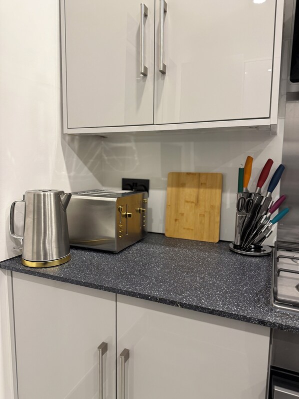 A kitchen with white cabinets, a toaster, a kettle, and a cutting board. - Cosy 2 Bedroom Flat in the Heart of Greenwich (London)