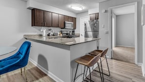 A modern kitchen with dark wooden cabinets, a granite countertop, a stainless steel refrigerator, and a built-in microwave. There are two bar stools and a blue chair. - Serene Waterfront 2BR  Poolside Living in Fort Lauderdale (Fort Lauderdale)