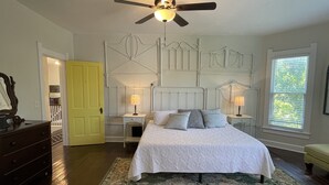 Ein Schlafzimmer mit einem Bett, zwei Nachttischen mit Lampen, einem Deckenventilator, einer gelben Tür und einem Fenster mit Jalousien.